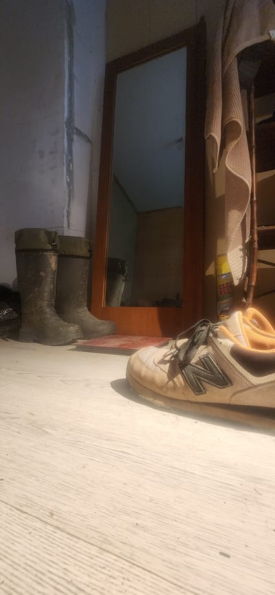 The image shows a pair of dark green rubber boots and a close-up of a New Balance sneaker. The boots are positioned on a light-colored, possibly wooden, floor. They appear to be outdoors or in an unfinished indoor space, as suggested by the visible concrete blocks and a wooden framed mirror in the background. The mirror reflects an empty room with light blue walls. Above the mirror, there's a glimpse of a rolled-up rug and a yellow can, possibly of cleaning spray. The lighting suggests it is daytime, with sunlight casting shadows. The scene appears to be utilitarian, possibly a garage or a storage area. There is no visible text in the image. The overall mood is functional and somewhat stark.