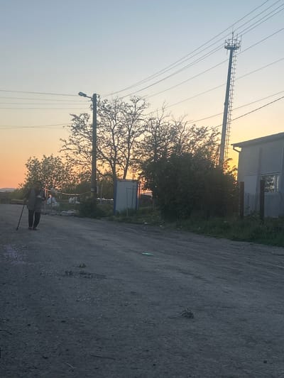 The scene is an exterior view of an unpaved road in Kalitinovo, Bulgaria. An individual wearing a jacket and dark pants stands on the road holding a walking stick. A metal pole with an attached streetlamp is visible near the individual, next to a bare tree. Behind the tree is a white structure with a blue trim. Further in the background is a building with a window and a power line tower. The sky is an even gradient of pale blue to orange, indicating sunset or sunrise. The road is asphalt with visible cracks.