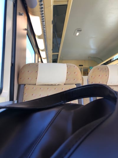 This image depicts the interior of a train carriage, presumably in the vicinity of Steinbourg, France. In the foreground, a dark, possibly black, bag with a prominent strap is visible, resting on what appears to be a seat or platform. The bag's material appears smooth, with visible stitching. Beyond the bag, two empty train seats are centered in the midground. These seats feature beige upholstery with a pattern of small, diagonal pink/red and light brown/gold dashes, and each is fitted with a clean white disposable headrest cover. The seat frames are grey.

To the left, a train window reveals an out-of-focus, brightly lit exterior. The ceiling of the carriage is light-colored, likely beige or off-white, and features multiple rectangular fluorescent light fixtures running lengthwise, providing illumination. A strip of small, circular vents or fasteners runs parallel to the lights along the ceiling. Overhead compartments are visible above the windows. Further down the aisle, more empty seats can be discerned, and a small, dark sign with light characters is visible above the seats in the distance. No individuals are present in the frame, and no specific actions or interactions are occurring. The scene conveys a clean, unoccupied train interior during daylight hours.