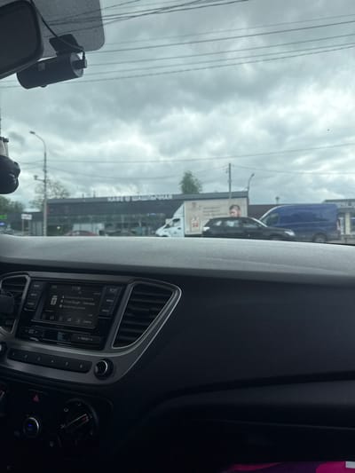 This image was taken from inside a car, looking out the windshield. The sky is overcast and appears to be cloudy. Outside the car, there is a large building with signage that includes the Russian words "КАФЕ ШАШЛЫЧНАЯ" (Cafe Shashlychnaya), indicating it is a cafe that serves shashlik. There are also vehicles visible outside, including a blue van and a dark-colored car. A dashcam is mounted on the windshield. The car's dashboard is visible, featuring a multimedia system displaying the time as 17:08 and information about a song. The overall setting appears to be a street in Molokovo, Russia, likely during the daytime.