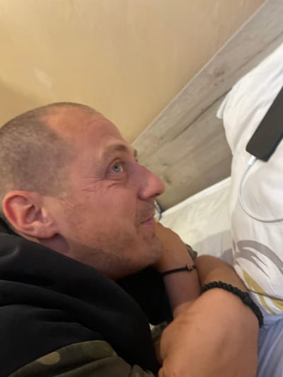 The image captures an adult man lying down in an indoor setting, possibly a bedroom or living area, in Gara Elin Pelin, Bulgaria. The shot is taken from an overhead perspective and rotated, showing the man's head and upper torso. He has very short, light-colored hair and light blue eyes, with a neutral, slightly contemplative expression. He wears a dark-colored top, possibly a hoodie, and a camouflage-patterned garment is visible on his right shoulder/arm. A black braided bracelet is on his right wrist, and a thinner black bracelet is on his left.
Next to him, resting on a white pillow or sheet, is a dark-colored smartphone with a white charging cable plugged into it. The background features a light beige wall or surface and wooden flooring or paneling. The man appears to be resting or looking towards something out of frame. The lighting suggests an indoor environment without strong natural light, providing no specific time-of-day indication. No text is visible in the image.