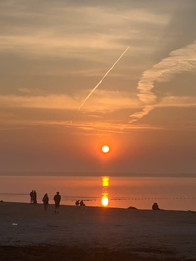The image depicts a serene sunset over a body of water, likely the Volga River, as viewed from the shore in Surakovo, Russia. The sky is painted in hues of orange and yellow, with wispy clouds and a prominent contrail from an aircraft streaking across the upper portion. The sun, a bright orb, is partially submerged on the horizon, casting a shimmering reflection on the calm water. On the sandy beach in the foreground, several silhouetted figures are visible. Some are walking along the shore, while others appear to be sitting or standing in small groups, enjoying the twilight. The scene is characterized by a peaceful ambiance, with the natural beauty of the sunset dominating the composition.