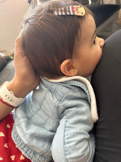 A baby is lying on an adult's arm, head resting on the adult's upper body. The baby is wearing a light blue denim jacket with a white hood lining and a red shirt with white heart patterns. A hair clip is in the baby's hair. The adult is wearing a white wristwatch with a red string bracelet. A dark-colored garment is visible behind the baby's head. The location is not discernible from the image.