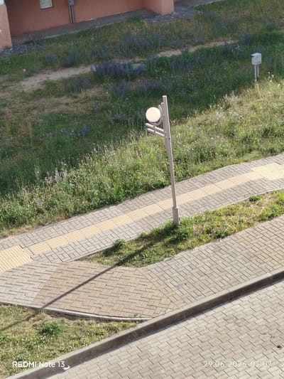 The image depicts an outdoor scene with a paved walkway and a grassy area. A lamppost with a spherical light fixture stands near the walkway. The walkway is composed of interlocking pavers with tactile paving strips, indicating it's likely a pedestrian path. Adjacent to the walkway is a grassy area with scattered wildflowers. In the background, a building with a reddish-brown facade is visible. The scene appears to be set in a park or urban green space. A small white electrical box on a post is also present in the grassy area. The timestamp "29.06.2026 07:07" and "REDMI Note 13" are present on the bottom of the image.