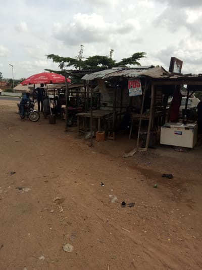 The image depicts a small roadside stall in Karshi, Nigeria. The stall is constructed from corrugated metal and wood, with a tarpaulin roof. A red umbrella branded with "Airtel" and "smartcash" stands near the front. Several individuals are present, some standing and observing, while one person sits on a motorcycle. Behind the stall, there are more motorcycles, wooden tables, and a white chest freezer. A sign on the stall reads "POS SERVICE". The ground is unpaved and dusty, with scattered debris, including plastic bags and wrappers. The sky is overcast.