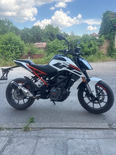 The image shows a white and black KTM motorcycle parked on a paved surface next to a sidewalk in Mihailovo, Bulgaria. Vegetation, including trees and overgrown plants, are visible in the background. The sky is blue with scattered white clouds. The motorcycle is the primary subject of the photo.