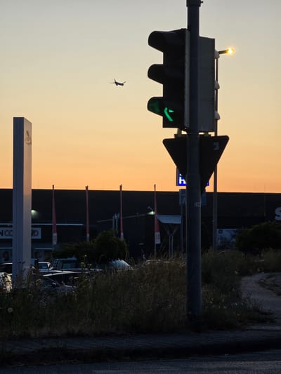 In Bischofsheim, Germany, at dusk, a traffic light with a green left-turn arrow is visible. An airplane is in flight in the orange-hued sky. A tall, white monument stands to the left, and a dark building with red flags is in the background. Several cars are parked or stopped near a grassy area. The scene appears to be near a commercial or industrial area.