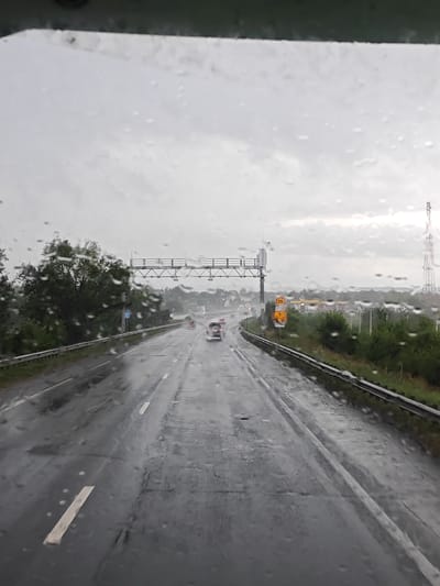 The image captures a view from inside a vehicle driving on a wet highway under an overcast sky. Raindrops are visible on the windshield, distorting the view of the road ahead. The highway appears to be multi-lane, with vehicles driving in the distance. Green trees and vegetation line the sides of the road, and a guardrail is visible. Several road signs are present, including a yellow circular sign with the number "25" inside, and a sign indicating a speed limit of "70". In the background, there are buildings that appear to be part of a town or service area, including a gas station with yellow canopies. A tall telecommunications tower is also visible in the distance. The overall atmosphere is gloomy due to the rainy weather. The scene is consistent with a highway in or near Belozerki, Russia.