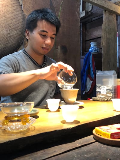 A man is seen pouring liquid from a glass sphere into a teacup at a wooden table. The man has short, dark hair and is wearing a grey t-shirt. The table is made of unfinished wood and is covered with various tea-related items, including a clear glass ashtray, several white teacups, a ceramic tea bowl, and some packaged tea. In the background, there are wooden beams and a woven mat. This scene appears to be taking place in a tea shop or a traditional tea ceremony setting, possibly in B'Lao, Vietnam, given the architectural style and cultural context often associated with tea rituals in the region.