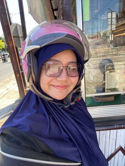 A woman wearing a dark blue hijab and a pink helmet with a clear visor is smiling at the camera. She is also wearing square-shaped glasses. Her face is partially obscured by the helmet and hijab. The woman appears to be standing outdoors, possibly on a street, as evidenced by the background which includes a road with a motorcycle in the distance and some vegetation. To her right, there is a glass window reflecting buildings and possibly the sky. A price list is visible on the window, suggesting the location might be a food stall or shop. The overall scene is brightly lit by sunlight. The location is identified as Sungai Paring, Indonesia.