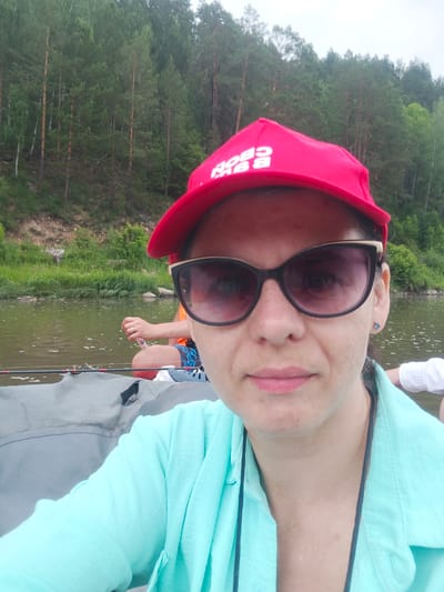A woman wearing a red baseball cap and sunglasses is seated in a boat on a river. The river is flanked by a densely wooded hillside. Another person, wearing a life vest and patterned shorts, is visible in the background. The woman is holding a fishing rod.