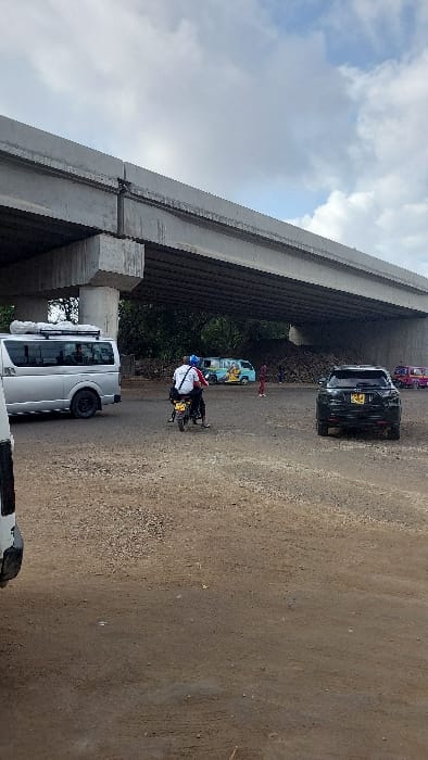 The scene depicts an unpaved, irregular dirt and gravel area beneath a multi-lane concrete overpass in Ziwa La Ng'ombe ward, Kenya. The sky above is partially overcast with white and grey clouds. A silver Toyota Hiace-style van with a black roof rack is positioned on the left, partially obscured by an object off-frame. In the mid-ground, a dark-colored motorcycle carries two individuals, both wearing helmets, traveling away from the camera. The rider wears a white collared shirt with a logo and dark trousers; the passenger wears a red shirt and dark trousers. To the right, a stationary black SUV displays a yellow Kenyan license plate. Underneath the concrete overpass, on the left, a light blue van with yellow accents (characteristic of a Kenyan 'matatu') is parked. Adjacent to it and further into the background, a substantial mound of dark, loose material, possibly soil or refuse, is present. Several distant figures are visible standing near this mound. On the far right, a pink and red three-wheeled auto-rickshaw (Tuk-tuk) is partially visible.