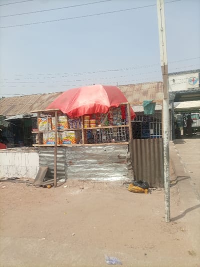 This image captures a roadside vending stall in Gulum, Nigeria, under a clear, light blue sky. The stall, constructed from a rudimentary wooden frame, chicken wire shelving, and a base of corrugated galvanized iron sheets, is centrally positioned. A large red umbrella provides shade over the merchandise.
The stall displays various packaged goods. On the upper left shelf, multiple yellow and red packages, some bearing the label "NATURE'S," are visible. The upper right shelf holds an assortment of smaller packaged items, including sachets and containers in red, yellow, and blue. A green cloth hangs near the right side of the stall.
The ground immediately surrounding the stall is unpaved, consisting of packed dirt with sparse debris, including a discarded yellow husk near the stall's base and a black plastic bag situated near a light-colored utility pole to the right. To the left of the stall, a low white wall and part of another structure with a corrugated metal roof are discernible. To the right, beyond the utility pole, a concrete sidewalk leads towards a building with an archway entrance. This building features a blue and white circular sign containing a white cross and a stylized 'E' or 'H', indicative of a medical or health facility. Overhead power lines are visible against the sky. No individuals are clearly engaged in specific actions or interactions in the immediate foreground; a blurry figure is faintly visible within the distant building's archway.