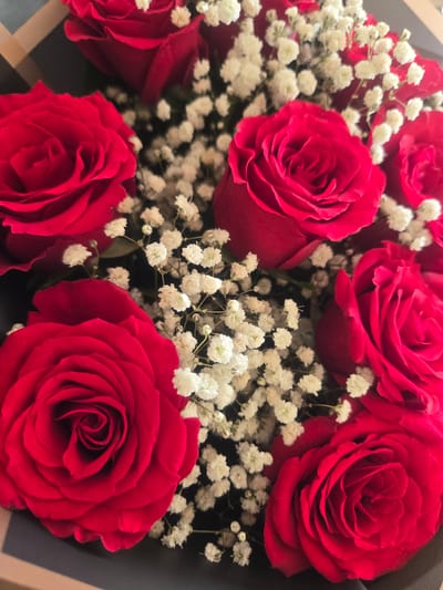 A close-up, overhead shot shows a bouquet of vibrant red roses intertwined with delicate white baby's breath flowers. The roses are in full bloom, with velvety petals forming intricate spiral patterns. The bouquet is arranged against a dark background, possibly a gift box or wrapping. The lighting is soft, highlighting the rich texture and deep color of the roses, creating a romantic and luxurious feel. The image is sharp and detailed, allowing for close examination of the floral arrangement. There are no people or discernible environmental features of Deira, United Arab Emirates present in the image.