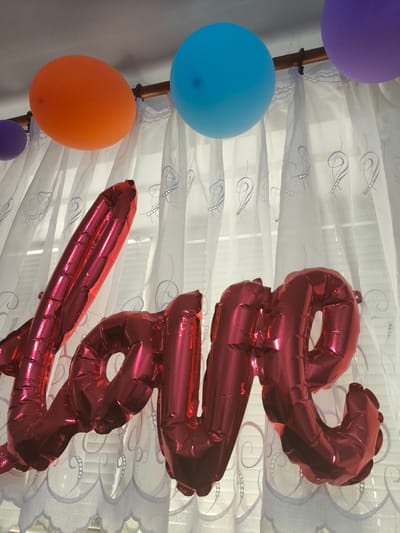 This media file showcases a decorative arrangement of balloons and foil letter balloons spelling out "LOVE". The "LOVE" balloons are large, metallic red, and individually shaped to form the letters. They are positioned prominently in the foreground.
In the background, several round balloons are visible, suspended from a wooden rod. These include an orange balloon, a bright blue balloon, and a purple balloon. Behind the balloons, a sheer white curtain with a decorative pattern is drawn. The pattern on the curtain appears to be a light-colored, swirling design. The overall impression is that of a festive or celebratory setting, possibly a room decorated for an event such as a birthday, anniversary, or Valentine's Day.
There is no visible text in the image, other than the word formed by the foil balloons. The lighting suggests it is daytime, but the specific time is not discernible. No people are visible in the frame. The setting appears to be indoors, with the curtain and rod suggesting a window treatment. The provided location context of Cricova, Moldova, does not offer any further visual cues within the image itself. The arrangement of balloons suggests an atmosphere of love, affection, or celebration.