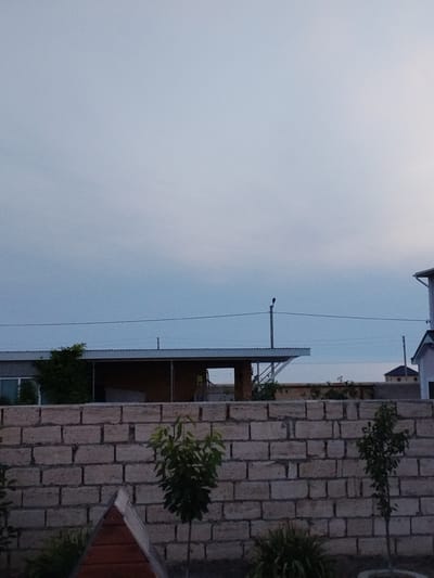 The image depicts an outdoor scene at dusk in Saiyn Şapağatov, Kazakhstan. A low, light-colored brick wall dominates the foreground, with sparse green foliage and a small, triangular wooden structure visible behind it. Beyond the wall, a long, low building with a metal roof is visible, partially obscured by a dense green bush on the left. Further in the background, a utility pole with a bird perched on top stands against a pale, overcast sky. To the right, a section of a modern building with a pitched roof is partially visible. The overall impression is one of a semi-developed or rural area with ongoing construction or development.