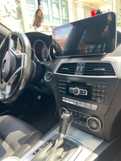 The image shows the interior of a Mercedes-Benz car. The dashboard features a large infotainment screen, climate control vents, a radio and navigation system, and a gear shifter. The steering wheel is visible on the left, and the leather seats are in the foreground. There are also decorative tassels hanging from the rearview mirror. The car appears to be parked outdoors, with sunlight filtering into the cabin. The exterior environment is partially visible through the windshield, suggesting an urban setting with buildings and possibly shops. There are no people visible in the car or in the immediate vicinity. The car's interior is clean and well-maintained.