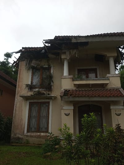 A two-story dilapidated building is shown. The roof is partially collapsed, revealing exposed rafters. The building's facade is faded and peeling, with plants growing on the windowsills. Two prominent pillars support a second-story balcony. Green vegetation obscures the base of the building. A partial view of another building is visible on the left. The scene conveys a sense of abandonment and disrepair.