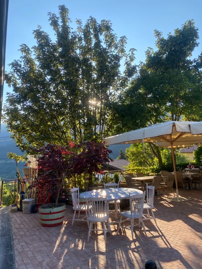 A sunny outdoor seating area with brick paving is set up for dining. A large, round white table with four matching white wooden chairs is positioned in the foreground, bathed in sunlight. Shadows from the table and chairs are cast across the brickwork. To the left of the table, a small red-leafed tree is planted in a wooden barrel decorated with red and white stripes. Behind the tree, a collection of gardening tools, including a broom and a pressure washer, leans against a stack of firewood. In the mid-ground, a large beige outdoor umbrella shades several tables and chairs, some of which are occupied by diners in the background. The seating area overlooks a scenic view of rolling hills and buildings, suggesting a location in or near the city of Caprino Veronese, Italy. Lush green trees with dappled sunlight filtering through their leaves dominate the background, and the sky is clear and blue.
