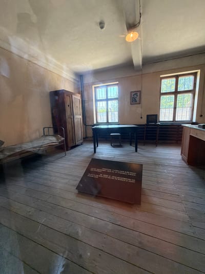 The image depicts a room furnished as it might have been in the Auschwitz-Birkenau concentration camp in Oświęcim, Poland. The room contains a simple wooden bed with a thin mattress, a tall wooden wardrobe, a desk with a chair and a small monitor, and two large, barred windows. The floor is made of wooden planks, and the walls are a pale, textured color. On the floor in the foreground, there is a dark, angled plaque with text in Polish and English. The lighting appears to be from a single overhead bulb and natural light from the windows. The overall impression is one of stark austerity and historical preservation.