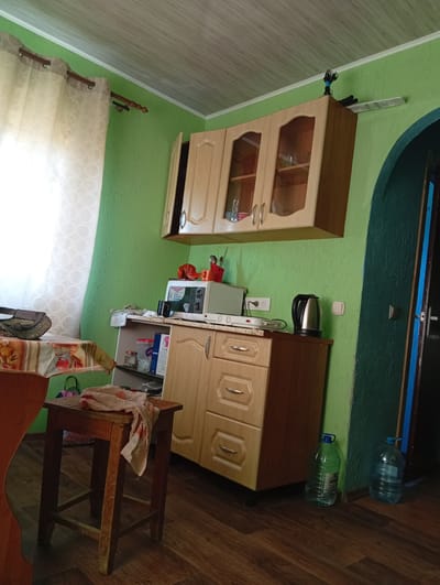 The image depicts an interior scene, likely a kitchen, in the Кліноцкі сельскі Савет, Belarus. The walls are painted a vibrant green with a textured finish. A wooden cabinet with glass-fronted doors is mounted on the upper part of the wall. Below it, a countertop holds a microwave, a kettle, and various containers. A wooden cabinet with drawers and doors is situated on the countertop. To the left, a table is covered with a patterned tablecloth, and a stool with a cloth draped over it stands nearby. A window with a sheer curtain is visible on the left side of the room. Two large plastic bottles of water are placed on the floor to the right. An open doorway, painted dark green with a blue trim, leads out of the room. The floor is covered with wooden planks. The ceiling is paneled. The overall impression is of a functional, somewhat cluttered domestic space. There are no people present in the image.