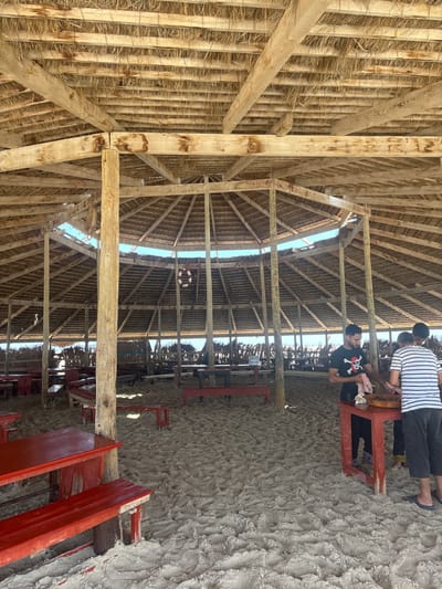 The image depicts a rustic open-air structure with a thatched roof supported by wooden beams and posts, situated on a sandy beach. The structure appears to be a restaurant or gathering space, with numerous red picnic tables and benches scattered across the sand. In the background, a fence-like barrier constructed from reeds or similar material stretches along the horizon.

In the foreground, three individuals are visible. Two men stand behind a red table, engaged in an activity that appears to be food preparation or serving. One man, wearing a black t-shirt with a skull and crossbones design, is holding a pink cloth. The other man, wearing a striped t-shirt and dark shorts, is looking down at the table. A third person, partially visible, stands behind the man in the striped shirt.

The environment is characterized by bright sunlight, casting shadows on the sand and the structure. The overall impression is of a casual, beachfront establishment. The location in Ghizen, Tunisia, suggests a coastal setting typical of the region.