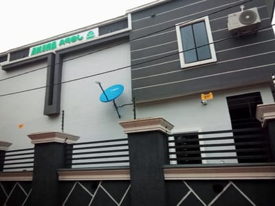 In Amokpara, Nigeria, the image captures the exterior of a modern, two-story building with a distinctive facade. The upper portion of the building features a dark gray, horizontally striped design, contrasting with a lighter gray section on the left. A prominent green sign with white lettering that reads "AMOKPARA ARENA" is visible on the left side of the upper facade, along with a small green logo. A single window with dark tinted glass and a white frame is centrally located on the upper right section. An air conditioning unit is mounted on the wall above the window.
Below the upper section, the building has a lighter gray wall on the left, and a dark gray and white striped fence or railing runs across the middle section. Black, horizontal metal bars are visible behind the fence. A bright blue satellite dish is mounted on the wall to the left of the window. Two black pillars with light-colored decorative caps stand in the foreground, supporting a decorative railing. Electrical wires are strung across the building's facade. A small yellow sign and a security camera are affixed to the wall below the window. The lower part of the image shows a dark wall with a geometric white pattern. The sky is overcast and appears bright, suggesting a daytime scene. No people are visible in the image.