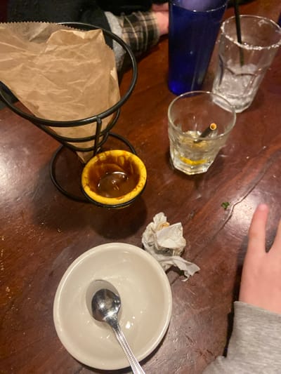 The image shows a wooden table in a restaurant. There is a brown paper bag in a wire holder, a small yellow bowl with brown liquid, a white plate with a spoon, a crumpled napkin, three glasses, and a hand on the right side of the frame. The glasses are empty and one has a straw in it. The hand is partially visible and appears to be reaching for the napkin. The coordinates provided (41.021, -73.635) indicate that the location is in the city of Greenwich, United States. The table is covered with a brown wooden surface. The scene appears to be a typical post-meal situation in a restaurant.