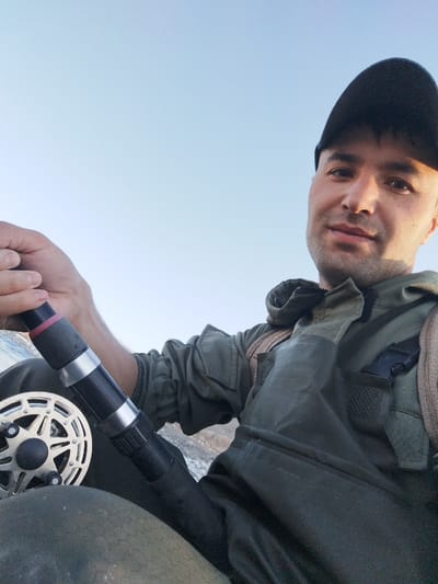 A single adult male is depicted in a close-up, upward-angled view against a clear, bright blue sky. He wears a black baseball cap and an olive-green, possibly waterproof, fishing suit or waders. A light brown strap, consistent with a backpack or chest harness, is visible over his right shoulder, secured by a black plastic buckle on the chest. The male subject, with short dark hair and light stubble, holds a fishing rod fitted with a silver-colored reel in his left hand, resting it across his lap. He looks directly into the camera with a neutral facial expression. The environment is an open outdoor setting during daytime. The location is in the city of Qoraqiya, Uzbekistan.
