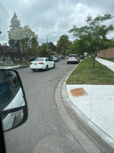 The image captures a residential street in Mississauga, Canada, under an overcast sky. A white Volkswagen sedan, license plate CHRZ939, is parked on the right side of the road, facing away from the viewer. To its right, a silver SUV is also parked. The street is lined with trees and houses, and a sidewalk is visible along the right side of the road. The foreground of the image shows a portion of a white vehicle's side mirror, reflecting the street and surrounding environment. The presence of traffic lights and street lamps suggests an urban or suburban setting. The overall scene depicts a typical day on a quiet street, with cars parked and trees lining the sidewalks.