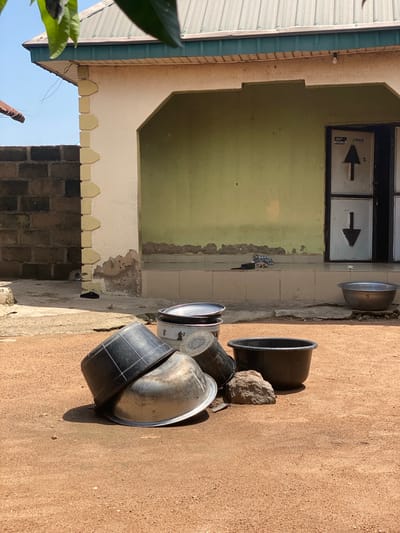 In Daniel, Nigeria, a sun-drenched outdoor scene unfolds on a sandy, reddish-brown ground. A light yellow stucco building with a green facade dominates the background, showing signs of wear with peeling paint near the base. The building features a textured green wall under a metal corrugated roof, accented by a green eave and trim. To the left, a dark brick wall stands adjacent to the building, its texture rough and weathered.
In the foreground, a collection of household items is scattered. Two large black plastic tubs are prominently displayed, one on top of the other, tilted at an angle. A large, silver metal basin is nestled between them, and another similar basin rests on a tiled step leading up to the building's entrance. A substantial, rough-hewn gray rock sits next to the black tubs.
The entrance to the building consists of two metal doors with diamond-patterned panels and a large black arrow pointing downwards on the right door. Above the entrance, a single light bulb is visible. Near the building's wall, a dark sandal and some folded fabric are placed on the tiled step, suggesting recent activity. The environment appears dry and arid, under a clear blue sky.