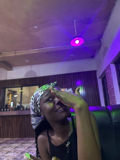 A young Black woman, wearing a silk headscarf and a black tank top with a delicate gold necklace, is captured in a close-up shot. She is looking upwards and to her right, with her right hand partially covering her face. The background features a wooden paneled wall and a glimpse of a dimly lit room with purple and green lighting. The ceiling shows exposed beams and a hanging lamp with a pink light. The woman's face is illuminated by the colored lights, casting a greenish hue on her cheek. She is seated on a black, leather-like surface, likely a couch or chair, with a patterned tiled floor visible at the bottom. A mobile phone with a bright green case is held in her left hand, partially visible at the lower left of the frame. The setting appears to be an indoor establishment, possibly a bar or lounge, with the presence of a bar counter visible through a glass partition in the background. The overall ambiance is dimly lit and vibrant due to the colored lighting.