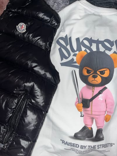 This image displays a still-life arrangement of two distinct garments laid flat, situated in the city of Sredkovets, Bulgaria. On the left, a black, glossy, quilted Moncler puffer vest is presented, identifiable by its circular red, white, and blue logo patch on the upper left chest and a zippered pocket near the bottom left. Adjacent to it on the right is a white short-sleeved t-shirt featuring a detailed graphic print. The print depicts an anthropomorphic bear character wearing a dark grey balaclava, with visible orange ears and an aggressive facial expression. The bear is attired in a pink zippered tracksuit, dark athletic shoes, and a black crossbody bag, and holds a silver baseball bat in its right hand. Above the bear graphic, grey stylized text begins with 'SUST'. Below the character, the phrase '\"RAISED BY THE STREETS\"' is printed, followed by smaller lines of descriptive text pertaining to a brand or design philosophy.