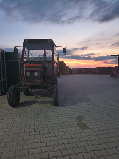 This is a full-shot, eye-level, outdoor photograph taken at dusk in Galonki, Poland. The image features a red tractor parked on a paved surface in front of stacks of pallets. The sky above is filled with dramatic clouds, tinged with the warm colors of the setting sun. To the left of the tractor, there are tall stacks of green material, likely construction or agricultural supplies. To the right, more stacks of wooden pallets are visible, extending into the distance. A simple roofed structure can be seen in the far right background. The tractor itself is the primary subject, with its front tires large and prominent. The paved ground is composed of interlocking concrete blocks, showing distinct patterns and shadows cast by the tractor and the evening light. No people are present in the image. The scene appears to be an outdoor storage or work area.