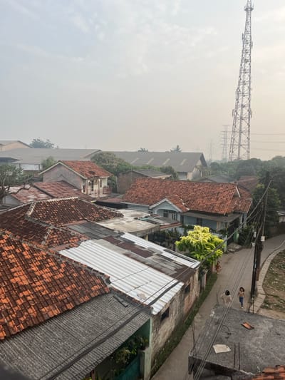 The image depicts a view of a densely populated urban residential area in Pasir Gadung, Indonesia, characterized by a network of houses with predominantly tiled roofs. In the background, a tall communication tower stands prominently, accompanied by power transmission lines and pylons, indicating the presence of significant infrastructure. The sky is hazy, suggesting potentially poor air quality or atmospheric conditions. A narrow road runs through the settlement, where two young girls are observed walking. The architecture of the houses appears varied, with some showing signs of age and others appearing more recently constructed. Vegetation, including trees and shrubs, is interspersed among the buildings. The overall impression is one of a working-class neighborhood with a mix of residential and industrial elements visible in the landscape.