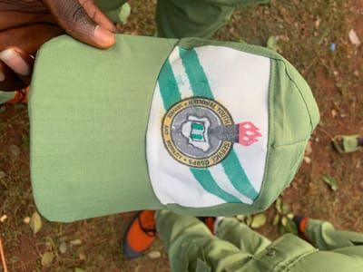 An overhead, close-up image depicts a light green baseball cap, held by a dark-skinned hand, in an outdoor setting in Kubi A, Nigeria. The cap features a light green brim and crown made of textured fabric. Its front panel is white, adorned with a prominent circular emblem of the National Youth Service Corps (NYSC). The emblem includes the text "NATIONAL YOUTH SERVICE CORPS" at the top, "SERVICE AND HUMILITY" at the bottom, a stylized map of Nigeria with a green rectangular object within, and a red rocket-like design to the right, all set against a background of green and white vertical stripes. In the blurred background, the lower legs and feet of a person are visible, wearing light green uniform trousers and orange and black athletic shoes, standing on reddish-brown earth with scattered dry grass and leaves.