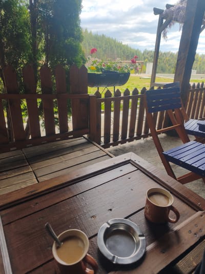 On a wooden table, two brown mugs of coffee and a metal ashtray are visible. Behind the table are two blue wooden chairs next to a wooden fence. Beyond the fence, there is greenery and some structures. The location is in Gorno Dryanovo, Bulgaria.