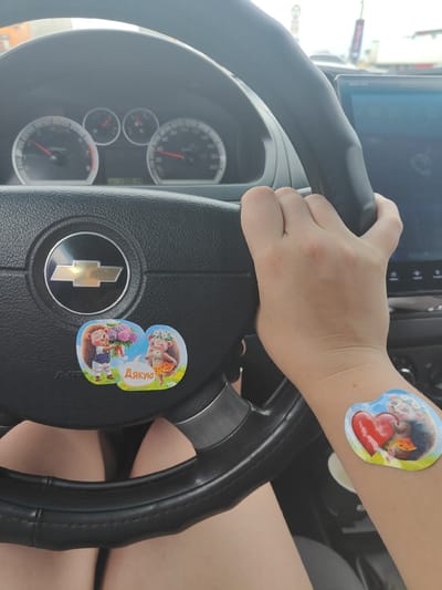 A person's hands grip the steering wheel of a car. A Chevrolet logo is visible on the steering wheel, along with two stickers depicting hedgehogs. One sticker shows a hedgehog holding a bouquet of flowers and the other shows a hedgehog wearing a flower crown, with the word "Дякую" (Thank you) in a speech bubble. Another sticker of a hedgehog holding a heart is visible on the person's forearm. The car's dashboard, including the speedometer and tachometer, is visible. The scene appears to be outdoors, possibly in a city, with traffic and buildings in the background. The location is Usatove, Ukraine.