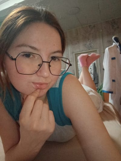 An indoor photograph depicts a female subject lying prone and facing the camera in a location identified as Старый Шагирт, Russia. The subject, with light brown hair, wears large, black-framed octagonal eyeglasses and a teal and grey tank top. Her right index finger is positioned on her lower lip. In the background, her left foot is visible, clad in a pink sock, with the leg bent at the knee. The wall behind her features light-colored, textured paneling. To the right, a white garment with blue trim, resembling a uniform, hangs on a fixture. The subject is situated on a surface covered with light-colored bedding.