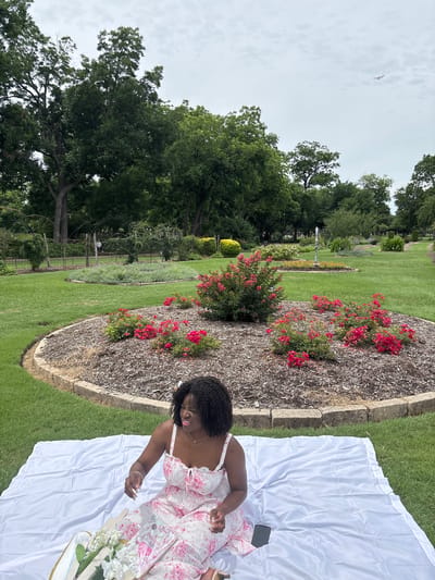 A high-angle, full outdoor shot depicts a woman reclining on a white sheet in a park setting. The woman has dark curly hair and wears a light-colored dress with a floral pattern. She is positioned on the right side of the frame and appears to be interacting with something out of view. To her left is a book and floral arrangement on the white sheet.
The setting is a grassy area in a garden or park with a bed of red flowers. The flower bed is edged with a low concrete barrier. Trees and other greenery are visible in the background, along with a wrought iron fence and other garden decorations. The sky above is overcast with a jet stream visible. The location appears to be Farmers Branch, United States of America.