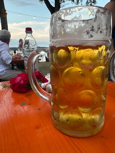 In Rimsting, Germany, on an outdoor table, a large stein of beer sits in the foreground, partially filled with golden beer and topped with foam. The stein has a textured glass design, and water droplets are visible on its exterior. To the left of the stein, on the orange wooden table, are two dark pink roses and some greenery. Behind the roses, a clear plastic bottle with a red cap stands upright. In the background, a woman with gray hair wearing a white top is seated at another table, facing away from the camera. Another person is visible behind her. Further in the background, lights are strung across the scene, and trees are present. A body of water, likely a lake, is visible beyond the tables, with a few people standing near it. The sky above is cloudy and shows the twilight hours.