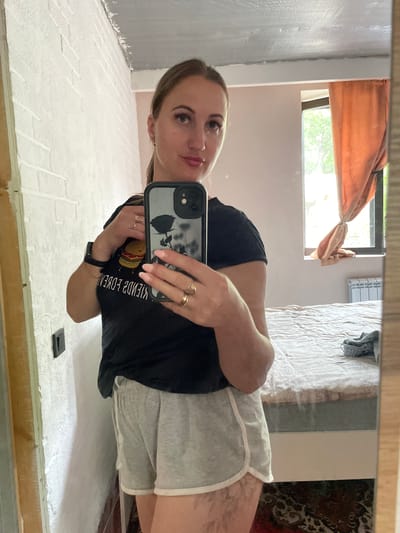 A woman is taking a mirror selfie. She is wearing a black t-shirt with a graphic of a hamburger and text on it, and grey shorts with white trim. She has light brown hair pulled back and is wearing rings on her fingers and a watch on her wrist. In the background, a bed with white bedding is visible, as is a window with orange curtains and trees outside. The room appears to be a bedroom. The image is oriented vertically, and the lighting suggests it was taken during the day. Some white brickwork is visible on the lower part of the mirror's frame.