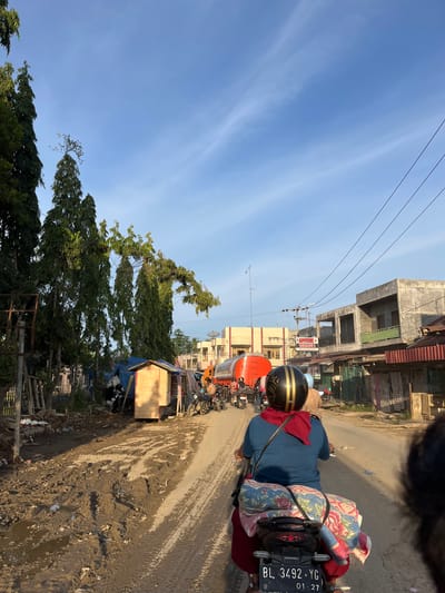 The image depicts a street scene in Alurcucur, Indonesia, characterized by a muddy, unpaved road. A person is riding a motorcycle in the foreground, from behind, wearing a blue shirt, red pants, and a black and gold helmet. A red scarf is draped around their neck, and a colorful blanket is secured to the back of the motorcycle. Other motorcycles and a large orange tanker truck are visible further down the road, along with several buildings lining the street. The sky is clear and blue with some wispy clouds. Vegetation, including tall trees and smaller plants, is present on the left side of the road. The overall impression is one of a developing or rural area with basic infrastructure.