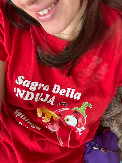 The image shows a person wearing a red t-shirt with the words "Sagra Della 'NDUJA" printed on it in white lettering. The 'NDUJA is a type of spicy spreadable salami originating in the Calabria region of Italy. The t-shirt also features a cartoon illustration of a red chili pepper with a human face. The person's face is partially visible in the top left corner of the image, along with a necklace. The image is taken indoors, as the background appears to be a fluffy brown blanket. The image is likely taken in Schaerbeek, Belgium, as the coordinates provided indicate a location in that city. There are no visible actions or interactions in the image. The subject appears to be wearing a red t-shirt as a statement about their love for 'NDUJA.