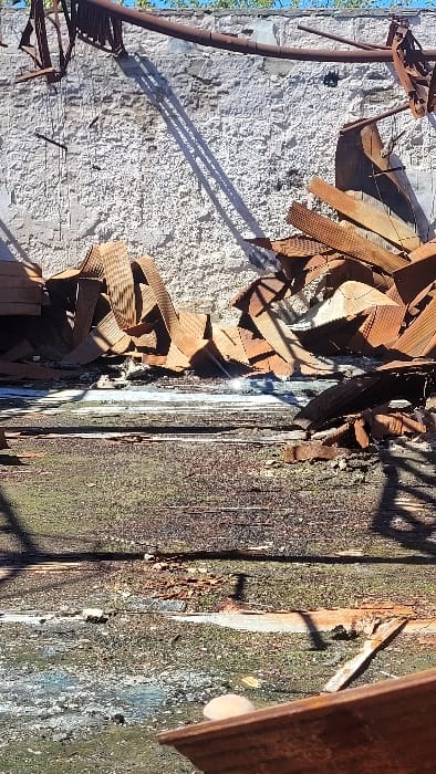 This image depicts an outdoor, derelict or industrial scene located in Zárate, Argentina, under bright, direct sunlight. The background features a tall, light-grey textured wall, exhibiting patches of discoloration and wear. Along the upper edge of the wall, a rusty horizontal metal pipe or beam is present, from which several twisted, rusty metal fragments extend. These elements cast sharp, elongated shadows diagonally across the wall, indicating strong illumination. At the base of the wall, a substantial pile of reddish-brown, severely corrugated and bent metal sheets, consistent with roofing or siding debris, is accumulated. Below this pile, an irregular band of mixed debris and shallow standing water is visible, with a prominent sun reflection. The foreground comprises a dark, heavily deteriorated ground surface, appearing to be decayed wooden planks covered in dark green moss, grime, and scattered detritus. Distinct horizontal lines suggest underlying wooden structure. Further elongated shadows traverse this foreground. In the lower right corner, a fragment of a dark, flat, possibly wooden or metal surface is partially visible, on which a single, light-colored, smooth, roundish object rests. No individuals are present in the scene.