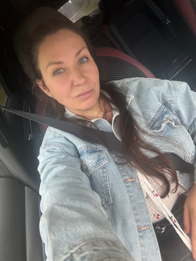 The media file is a close-up, rotated selfie of a woman sitting in what appears to be a car. She is wearing a light blue denim jacket over a white top with a cherry pattern. Her long, dark brown hair is visible. Her facial features, including her blue eyes, are prominent in the shot. A black seatbelt is visible across her body. The background suggests the interior of a vehicle, with glimpses of car seats and dark panels. The overall lighting indicates it's daytime, though no specific weather or time of day cues are strongly evident. The image conveys a casual and personal moment. There is no visible text in the image.