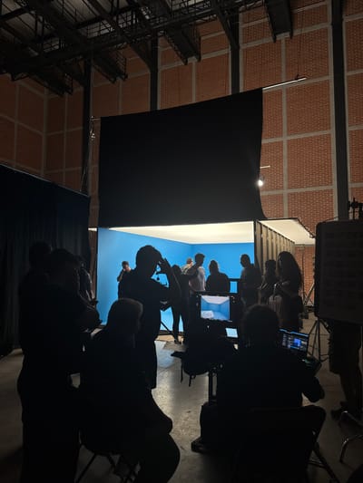 The scene depicts a film set in a large, industrial building with a brick ceiling, likely in Tlalnepantla, Mexico. A bright white screen with a blue image is visible, surrounded by black fabric. Silhouetted figures, some seated in chairs, are observing the screen. There's film equipment, including monitors and lighting, surrounding the screen and the individuals.