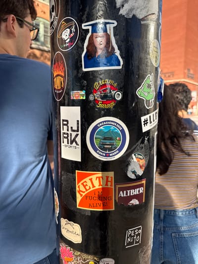 The image shows a black pole covered in an assortment of stickers. Text on the stickers includes "HUSA Ori booKIN AMIs RJ RK SPORTS Dreams Garage," "VACACIONES 2025 KOKIS MARIO VACACIONES FOREVER #LLG Travels Soypette," "KEITH I'M FUCKING ALIVE! ALTBIER," and "PESO Netol Nooo..! T 4M." Other stickers depict cartoon characters, automotive themes, and stylized designs. People stand or lie near the pole. The overall impression is a collection of personal expressions and interests displayed through sticker art.