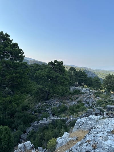 The image depicts a landscape view from a high vantage point, overlooking a valley filled with dense green trees. The foreground is dominated by large, weathered gray rocks, some covered in lichen, and dry, yellow grass. In the midground, the ruins of an ancient stone structure are visible, with many rectangular stone blocks scattered across the terrain, overgrown with vegetation. Further into the midground and background, rolling hills covered in a mix of trees and lighter green foliage recede into the distance, under a clear, pale blue sky. The scene appears to be a natural environment with historical remnants, likely located in the region of Seydikemer, Turkey, given the context. No people or specific actions are discernible.