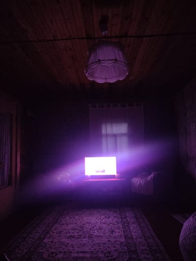 A dimly lit room, likely in Beryozovka, Russia, features a wooden paneled ceiling with a white fabric lampshade hanging from the center. A glowing television screen emits a bright purple light, casting a haze across the room and illuminating a patterned rug on the floor. A window with sheer curtains is visible behind the television. The overall atmosphere is dark and mysterious, with hints of furniture and personal items suggesting a living space.